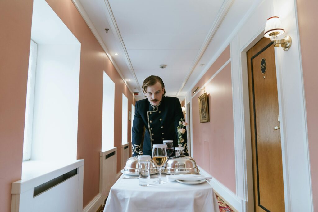 Bellboy in uniform presents room service in a hotel corridor setting.